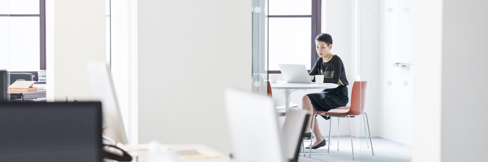 Zoomed out image of a woman in an office sitting at a desk and using a laptop