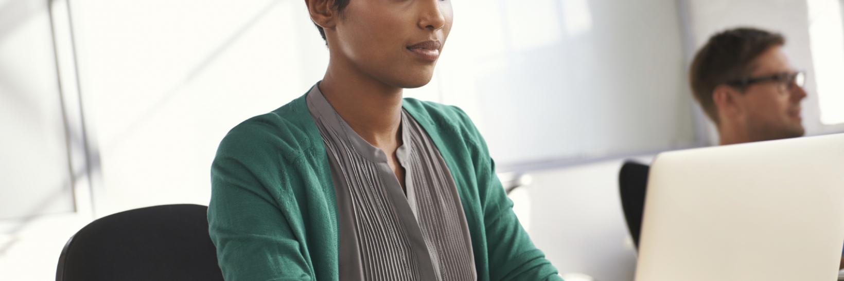 A woman typing on a silver lap top at her desk, with a co-worker in the backdrop