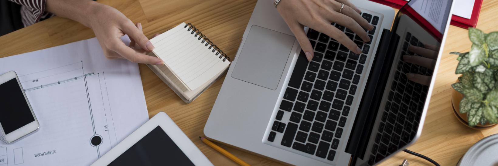 aerial view of a woman at laptop with a tablet on her desk and a note pad. 