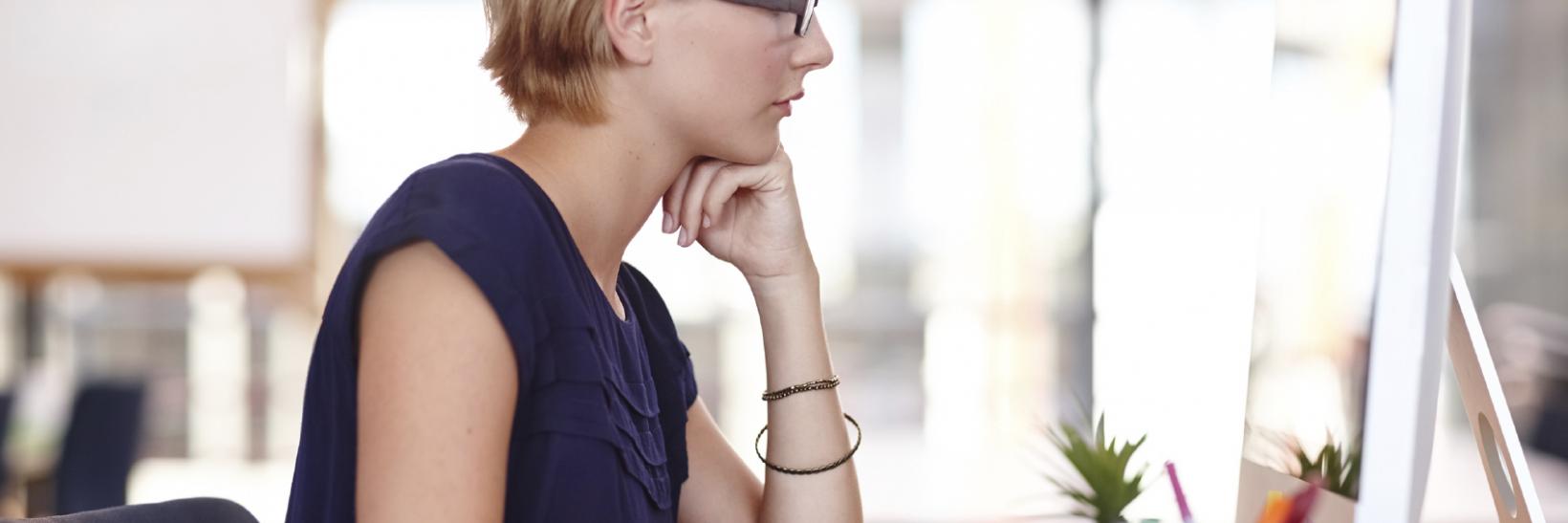 Blonde white woman sitting in front of a desk, on a computer. 