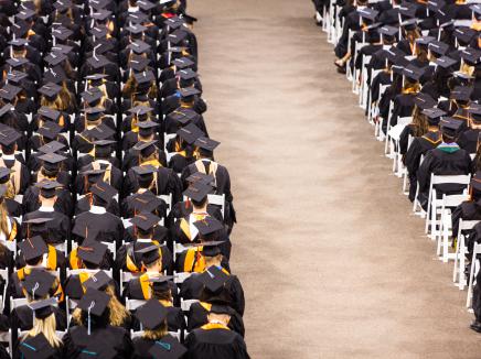 Room with rows of chairs and students sitting wearing black graduation caps and gowns