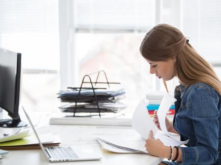 Woman sits a desk looking at a laptop and reviewing papers