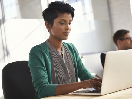 A woman typing on a silver lap top at her desk, with a co-worker in the backdrop 