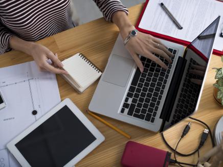 aerial view of a woman at laptop with a tablet on her desk and a note pad. 