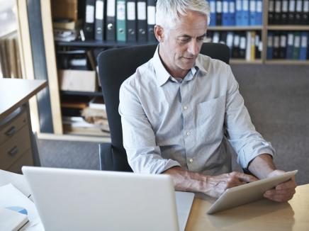 Man sitting at desk in front of a lap top, using his tablet