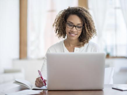 Young woman writing while looking at an open laptop computer