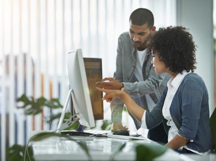 Man and woman looking at and pointing to two desktop computer monitors.