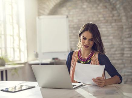 woman sitting at desk with her laptop open, reading a piece of paper